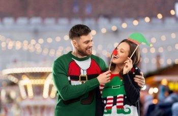 people and holidays concept - happy couple in sweaters posing with party props over christmas market background. couple in sweaters with christmas party props