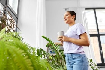 people, housekeeping and plants care concept - happy african american woman with houseplants drinking coffee and looking out window at home. woman with coffee looking out window
