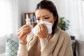 health, cold and people concept - sick young asian woman in blanket with nasal spray medicine blowing nose to paper tissue at home. sick asian woman with nasal spray blowing nose