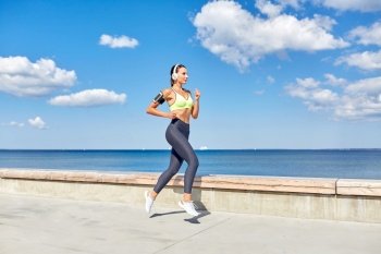fitness, sport and healthy lifestyle concept - young woman with headphones and smartphone in armband running at seaside. woman with headphones and smartphone running