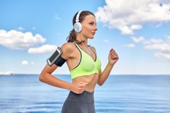 fitness, sport and healthy lifestyle concept - young woman with headphones and smartphone in armband running at seaside. woman with headphones and smartphone running