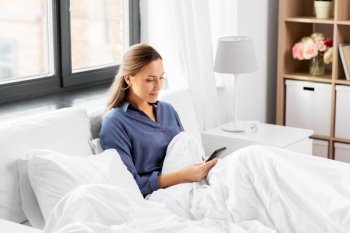 technology, internet, communication and people concept - happy young woman texting on smartphone in bed at home in morning. young woman with smartphone in bed in morning