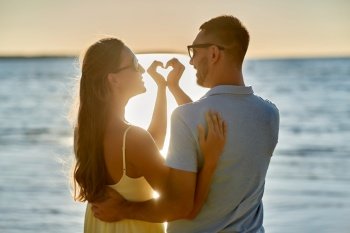 leisure, relationships and people concept - happy couple in sunglasses hugging and showing hand heart gesture on summer beach. happy couple showing hand heart on summer beach