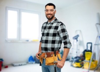 repair, construction and work concept - happy smiling male worker or builder in goggles with tool belt over room with building equipment background. happy male worker or builder with tool belt