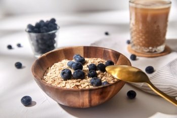 food, healthy eating and breakfast concept - oatmeal in wooden bowl with blueberries and spoon, glass of ice coffee on cork drink coaster and kitchen towel. oatmeal with blueberries, spoon and coffee