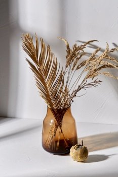 home improvement and decoration concept - still life of decorative dried flowers in brown glass vase and pumpkin dropping shadows on white surface. decorative dried flowers in glass vase and pumpkin