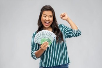 people, ethnicity and portrait concept - happy asian young woman holding hundreds of euro money banknotes celebrating success over grey background. happy asian woman with money celebrating success