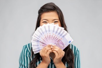people, ethnicity and portrait concept - happy asian young woman hiding her face behind hundreds of euro money banknotes over grey background. asian woman with hundred euro money banknotes
