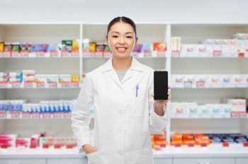 medicine, profession and healthcare concept - happy smiling asian female pharmacist or doctor with stethoscope showing smartphone over pharmacy background. asian pharmacist with smartphone at pharmacy