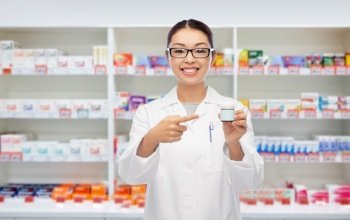 medicine, profession and healthcare concept - happy smiling asian female pharmacist or doctor holding jar of pills over pharmacy background. asian female pharmacist or doctor with medicine