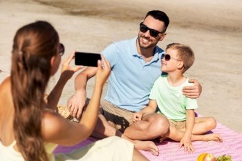 family, leisure and people concept - happy mother with smartphone photographing father and son on summer beach. family with smartphone photographing on beach