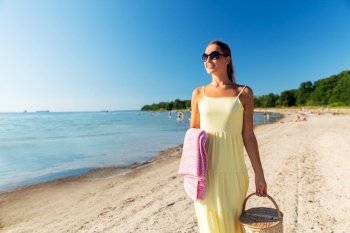 leisure, relationships and people concept - happy smiling woman in sunglasses with picnic basket and blanket walking along summer beach. happy woman with picnic basket walking along beach
