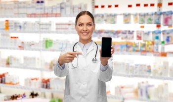 medicine, profession and healthcare concept - happy smiling female doctor or nurse with stethoscope showing smartphone over pharmacy background. happy female doctor or nurse with smartphone