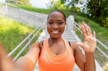 fitness, sport and technology concept - happy smiling young african american woman with wireless earphones listening to music, taking selfie and waving hand outdoors. happy african woman with earphones taking selfie