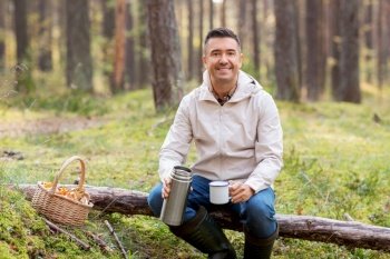 picking season and leisure people concept - smiling man with wicker basket of mushrooms sitting on fallen tree and drinking hot tea in autumn forest. man with basket of mushrooms drinks tea in forest