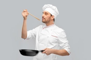cooking, culinary and people concept - happy smiling male chef in jacket with frying pan and spoon tasting food over grey background. happy male chef with frying pan tasting food