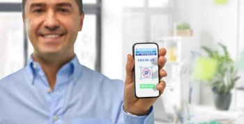 technology and health care concept - close up of smiling man holding and showing smartphone with international certificate of vaccination on screen over office background. man with certificate of vaccination on smartphone