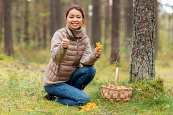 picking season, leisure and people concept - young asian woman with basket and chanterelle mushroom showing thumbs up in autumn forest. woman with mushrooms showing thumbs up in forest