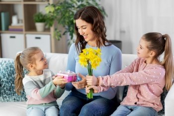 people, family and holidays concept - two daughters giving daffodil flowers and birthday present to happy mother at home. daughters giving flowers and gift to happy mother