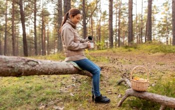 picking season, leisure and people concept - young asian woman with mushrooms in basket and thermos drinking tea in autumn forest. asian woman with mushrooms drinking tea in forest