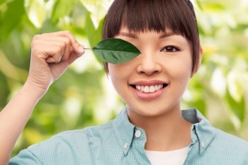 eco living, environment and sustainability concept - portrait of happy smiling young asian woman in turquoise shirt holding green leaf over natural background. smiling asian woman holding green leaf