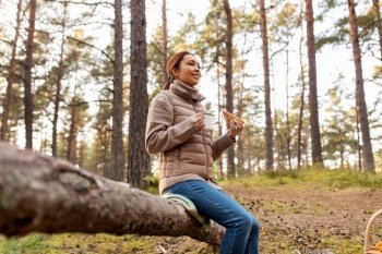 picking season, leisure and people concept - young asian woman with mushrooms in basket drinking tea and eating sandwich in autumn forest. woman with mushrooms drinks tea and eats in forest