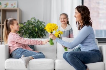 people, family and holidays concept - two daughters giving daffodil flowers to happy mother at home. daughters giving daffodil flowers to happy mother