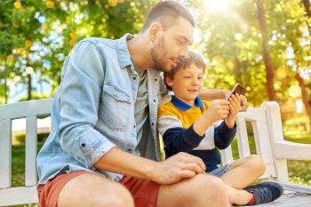 family, fatherhood and technology concept - happy father and little son with smartphone sitting on bench at summer park. father and son with smartphone at park