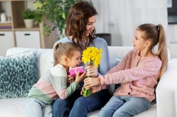 people, family and holidays concept - two daughters giving daffodil flowers and birthday present to happy mother at home. daughters giving flowers and gift to happy mother