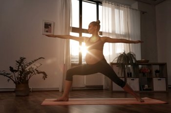 sport, fitness and people concept - happy pregnant woman doing yoga warrior pose at home over sunshine. pregnant woman doing yoga at home over sunshine