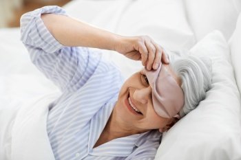 old age and people concept - happy smiling senior woman with eye sleeping mask in bed at home bedroom. senior woman with eye sleeping mask in bed at home