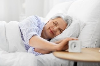 old age and people concept - happy smiling senior woman with alarm clock lying in bed at home bedroom. happy senior woman with alarm clock in bed at home