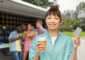 people and drinks concept - happy young asian woman drinking takeaway coffee from paper cup over food truck background. happy asian woman drinking coffee over food truck