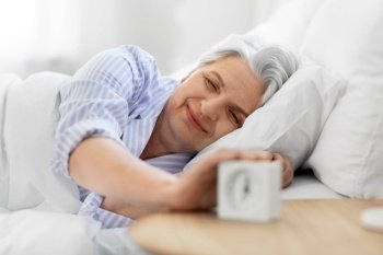 old age and people concept - happy smiling senior woman with alarm clock lying in bed at home bedroom. happy senior woman with alarm clock in bed at home