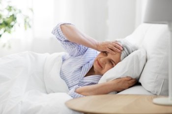 health, old age and people concept - senior woman in pajamas suffering from headache lying in bed at home bedroom. senior woman with headache in bed at home