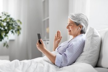 technology, old age and people concept - senior woman with smartphone and wireless earphones having video call in bed at home bedroom. senior woman with phone having video call in bed