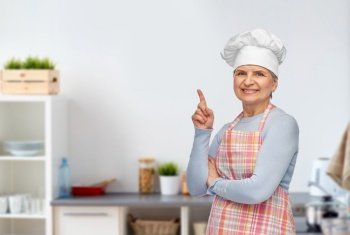 cooking, culinary and old people concept - portrait of smiling senior woman or chef in toque in apron pointing finger up over home kitchen background. senior woman or chef pointing finger up at kitchen