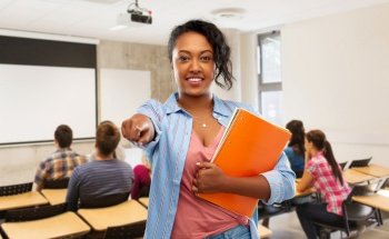 education, school and people concept - happy african american young student woman with notebooks pointing finger to camera over classroom background. african american student woman with notebooks