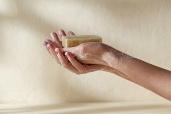 natural cosmetics, hygiene and beauty concept - foamy hands holding bar of craft soap on beige background. foamy hands holding bar of craft soap