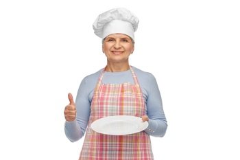 cooking, culinary and old people concept - portrait of smiling senior woman or chef in toque in kitchen apron holding empty plate showing thumbs up over white background. smiling senior woman or chef holding empty plate