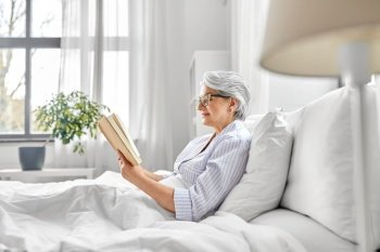 old age, leisure and people concept - happy smiling senior woman in glasses reading book in bed at home bedroom. old woman in glasses reading book in bed at home