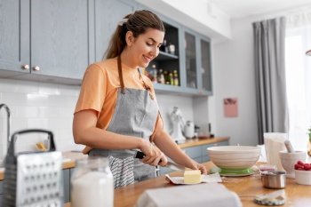 culinary, baking and people concept - happy smiling young woman cooking food on kitchen at home cutting butter with knife. woman cooking food and baking on kitchen at home