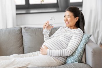 pregnancy, rest, people and expectation concept - happy smiling pregnant asian woman sitting on sofa at home and drinking water from reusable glass bottle. pregnant woman with water in glass bottle at home