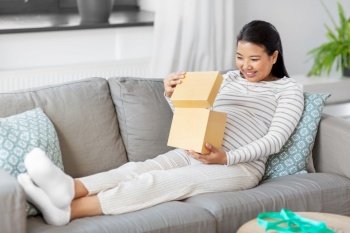 pregnancy, holidays and people concept - happy smiling pregnant asian woman opening gift box sitting on sofa at home. happy pregnant woman opening gift box at home
