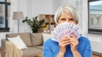 savings, finances and people concept - senior woman hiding her face behind fan of five hundred euro money banknotes over home living room background. senior woman with hundreds of euro money banknotes