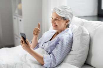 old age, technology and people concept - happy smiling senior woman in pajamas using smartphone sitting in bed and having video call and showing thumbs up at home bedroom. senior woman with phone having video call in bed