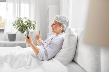technology, old age and people concept - senior woman with smartphone and wireless earphones having video call in bed at home bedroom. senior woman with phone having video call in bed