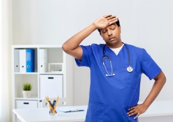 healthcare, profession and medicine concept - stressed indian doctor or male nurse in blue uniform over medical office at hospital background. stressed doctor or male nurse in blue uniform