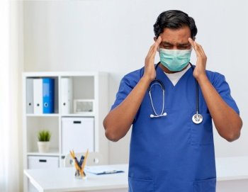 healthcare, profession and medicine concept - stressed indian doctor or male nurse in blue uniform and face protective medical mask having headache over medical office at hospital background. stressed indian doctor in mask having headache