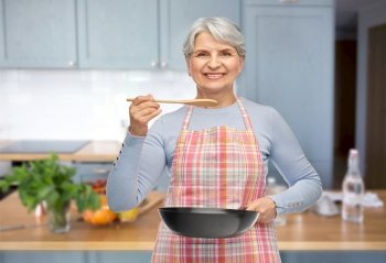 cooking, culinary and old people concept - portrait of smiling senior woman in apron with frying pan and spoon tasting food over kitchen background. smiling senior woman in apron with frying pan